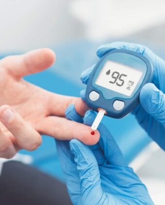 A high-quality, professional photograph focusing on blood sugar control, featuring a close-up of a healthcare provider's hands in blue clinical gloves performing a glucose test on an Indian patient's finger